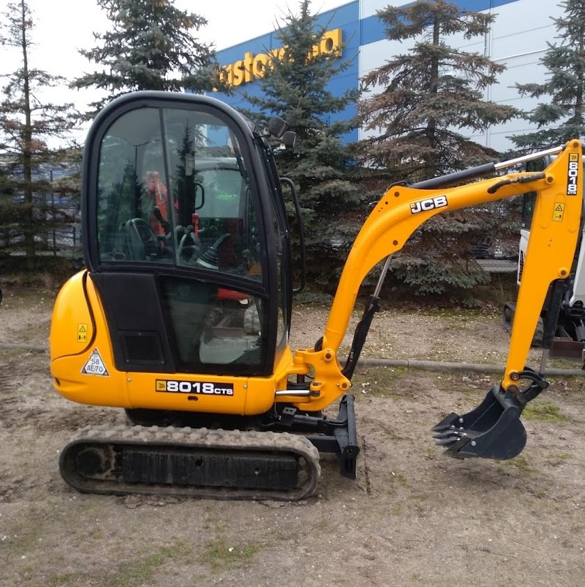 Żółta mini koparka JCB 8018 CTS na ziemi, z widocznym logiem JCB, na tle budynku Castorama i kilku choinek.