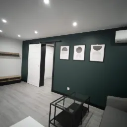 Nowoczesne wnętrze pokoju po remoncie: ciemnozielona ściana z trzema abstrakcyjnymi obrazami, szara podłoga z panelami, przesuwne białe drzwi, czarny stolik kawowy i klimatyzator.