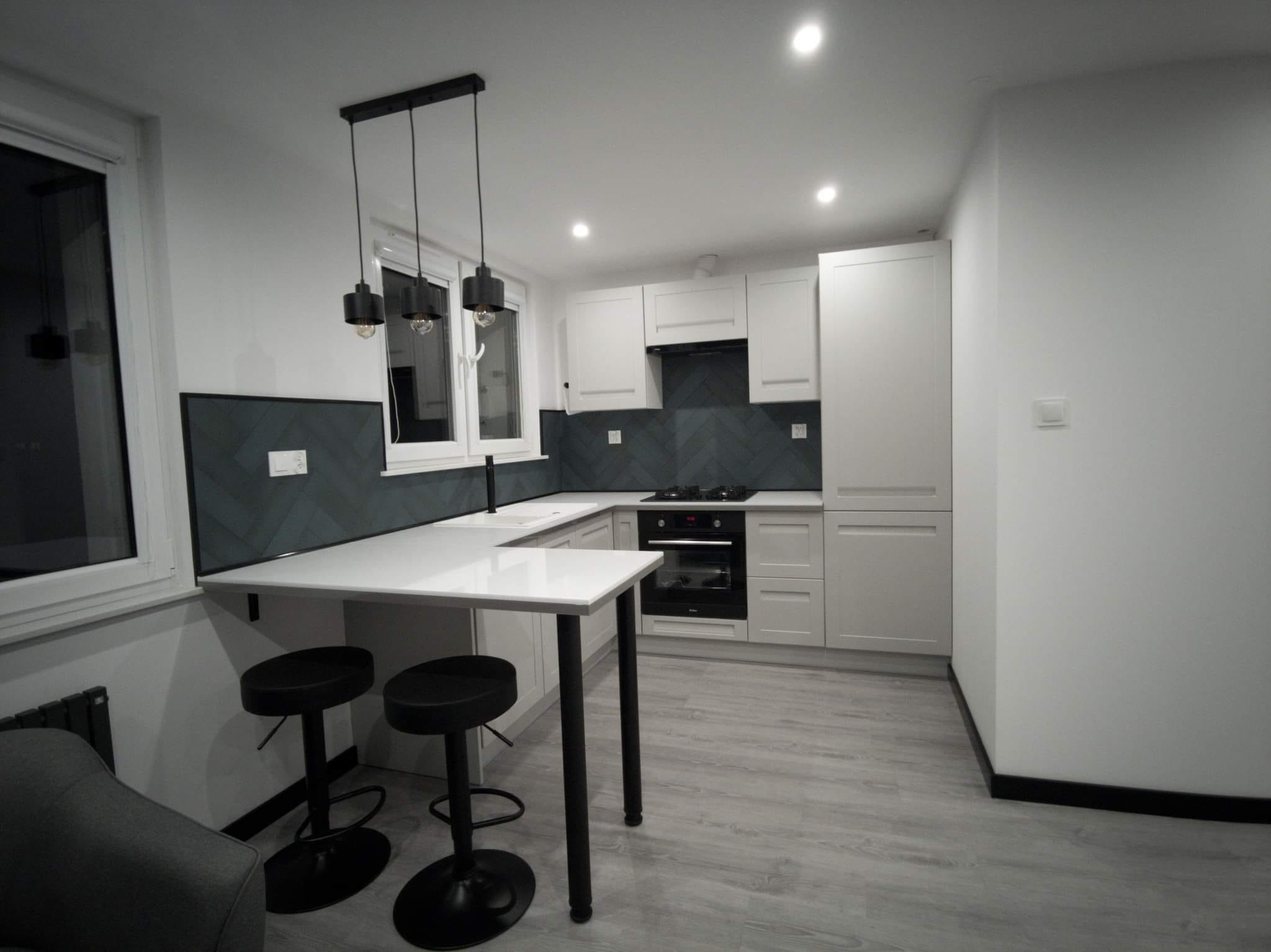 Nowoczesna kuchnia w odcieniach szarości i bieli, z czarnymi akcentami w postaci stołków barowych i lamp wiszących nad blatem. Ściana nad blatem wykończona płytkami ułożonymi w jodełkę.