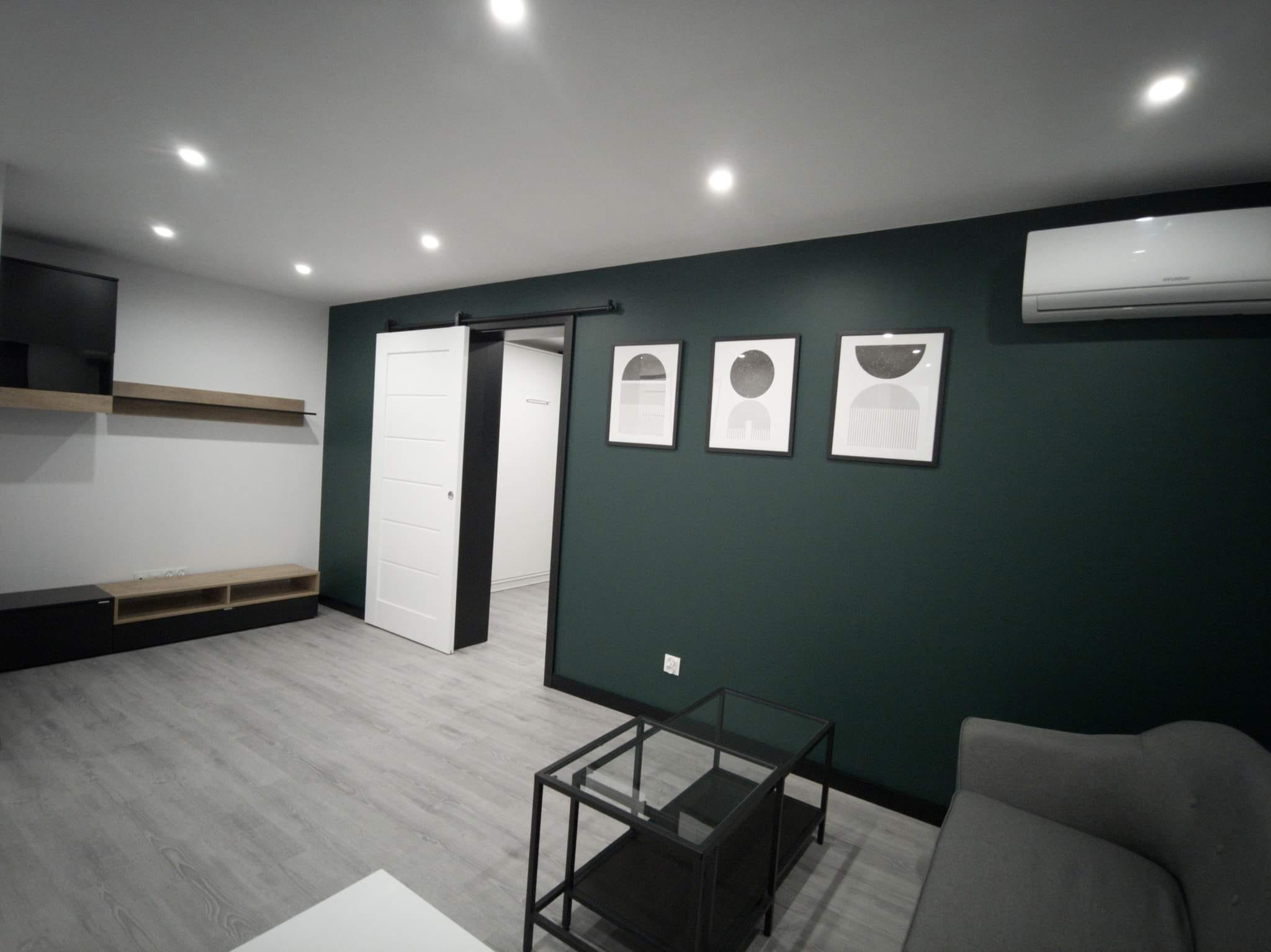 Nowoczesne wnętrze pokoju po remoncie: ciemnozielona ściana z trzema abstrakcyjnymi obrazami, szara podłoga z panelami, przesuwne białe drzwi, czarny stolik kawowy i klimatyzator.