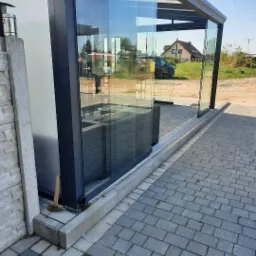 Zadaszony taras z przesuwnymi szklanymi ścianami, aluminiowa konstrukcja malowana proszkowo, widoczny młotek oparty o podstawę konstrukcji.