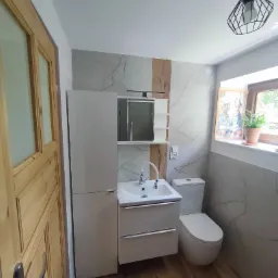 Nowoczesna łazienka po remoncie: biała umywalka z szafką, toaleta, marmurowe płytki z drewnianym akcentem, drewniane drzwi i designerska lampa.