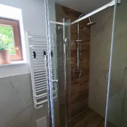 Nowoczesna kabina prysznicowa z chromowanymi okuciami, drewnopodobne płytki, grzejnik drabinkowy i okno z widokiem na zieleń.