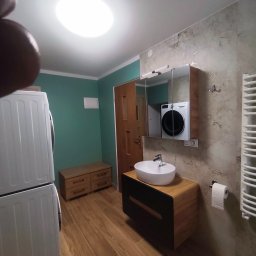 PE-ŁKA usługi budowlane - Jasna łazienka z drewnianymi akcentami: umywalka na szafce, pralka w lustrze, grzejnik drabinkowy i płytki imitujące marmur. Nowoczesny design z nutą naturalności.