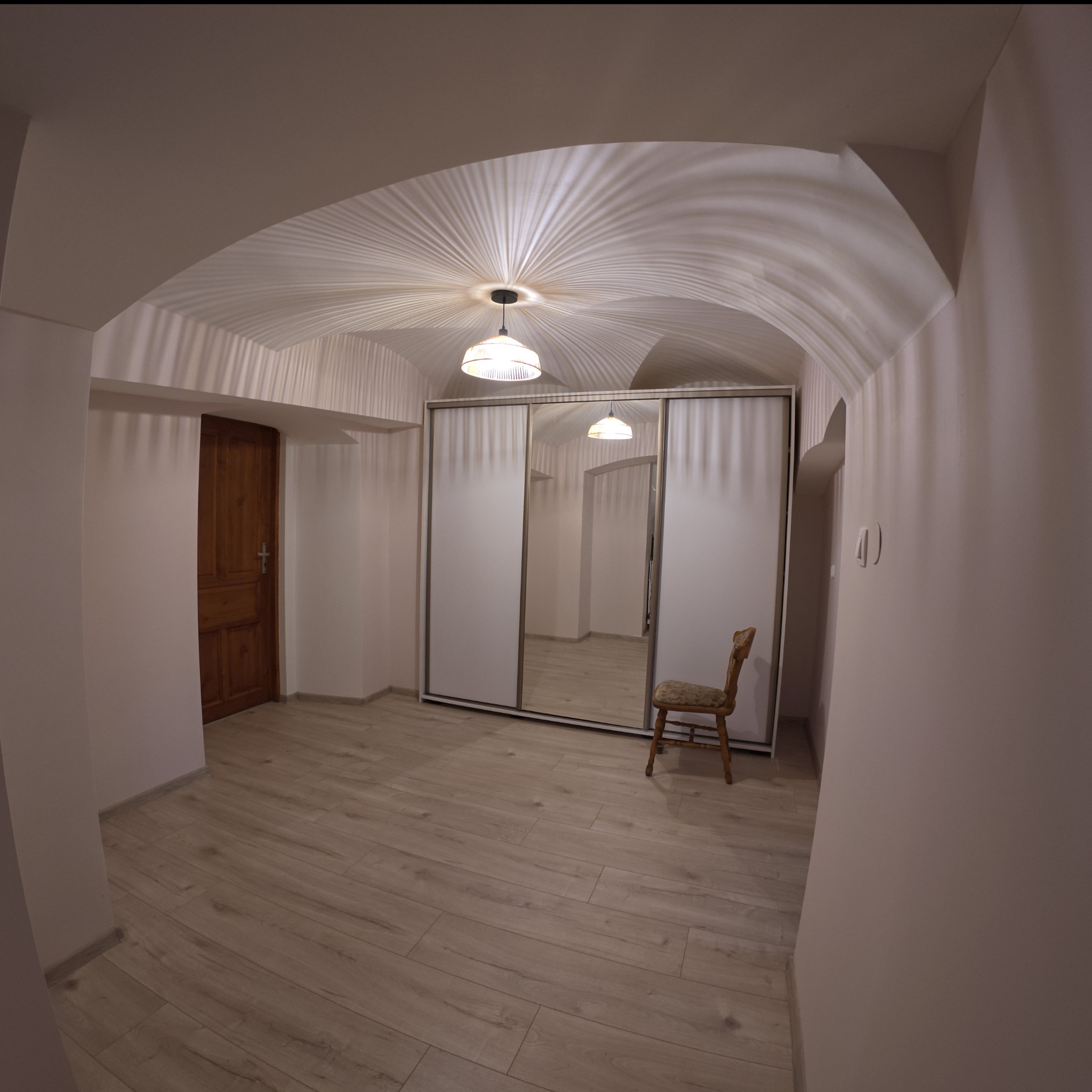 Jasne wnętrze z drewnianą podłogą, przesuwne drzwi szafy z lustrami, drewniane drzwi wejściowe oraz krzesło z tapicerowanym siedziskiem. Sufit z łukowym sklepieniem i lampą generującą promieniowe...