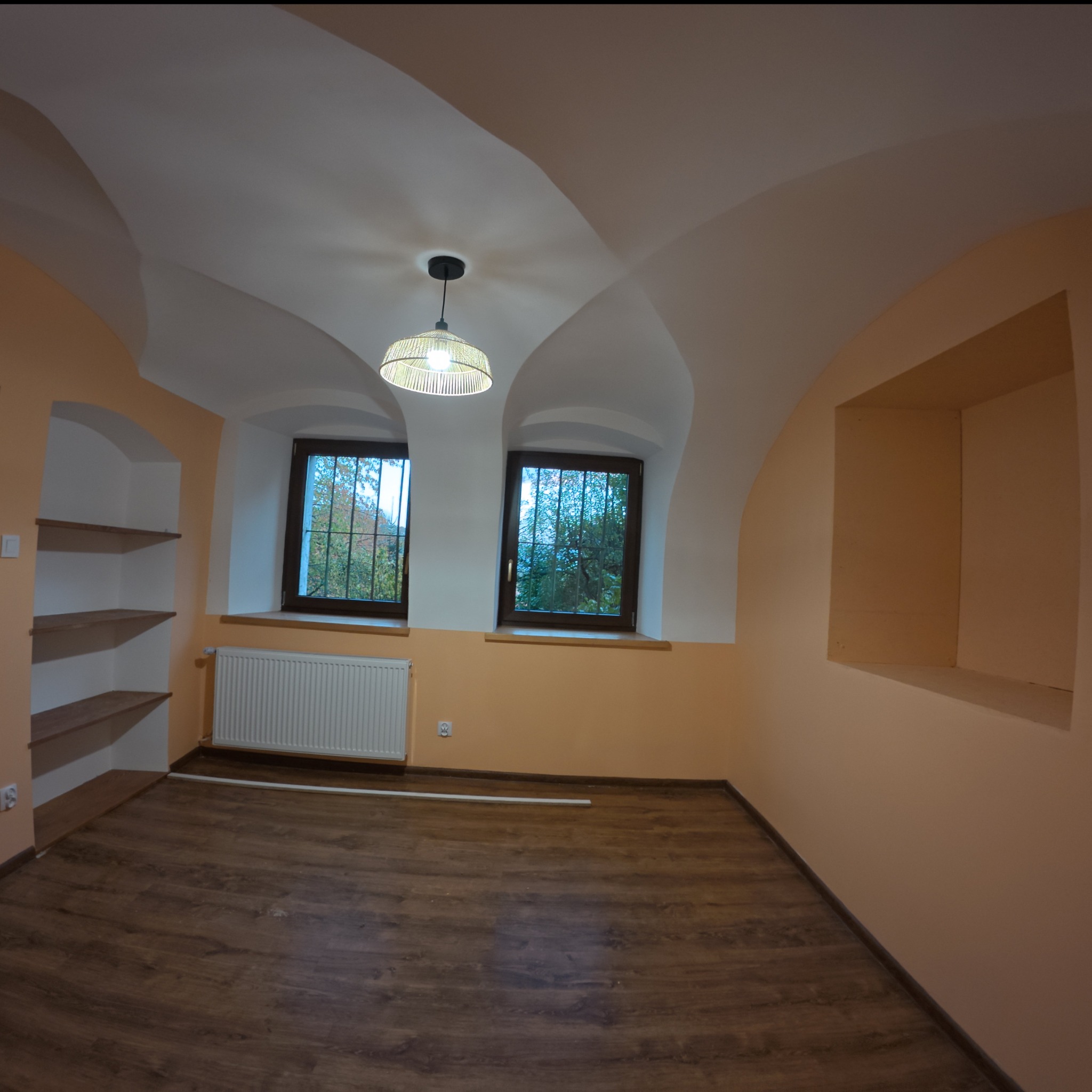 Pusty pokój po remoncie z panelami podłogowymi, dwoma oknami z kratami, grzejnikiem, wnęką na ścianie i półkami w ścianie, oświetlony lampą wiszącą z abażurem.