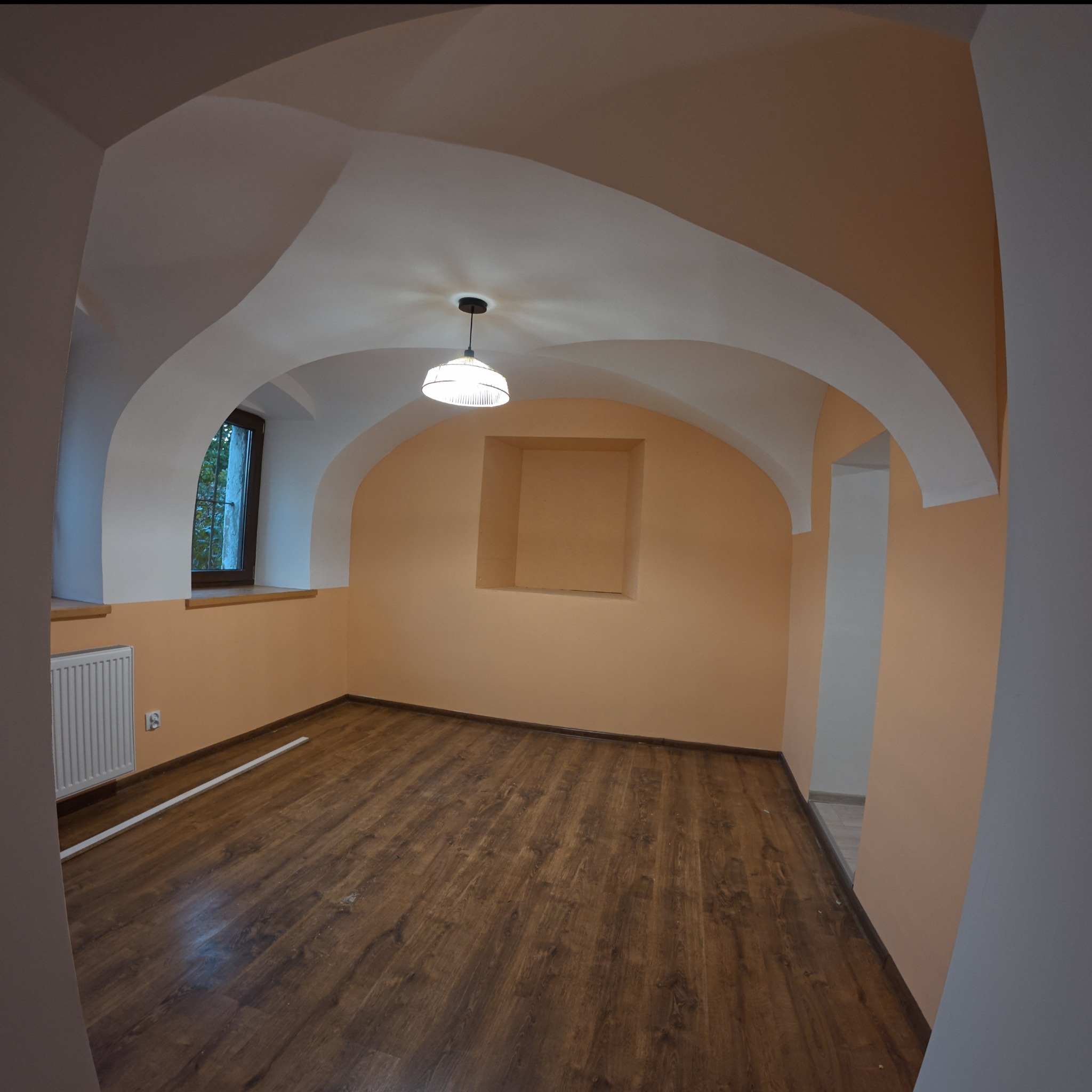 Pomalowane na brzoskwiniowo wnętrze pokoju z drewnianą podłogą, białymi sklepieniami i oknem z widokiem na zieleń, oświetlone wiszącą lampą z kloszem.