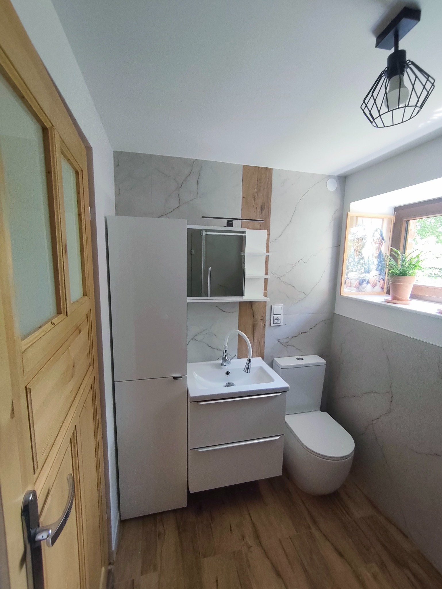 Nowoczesna łazienka po remoncie: biała umywalka z szafką, toaleta, marmurowe płytki z drewnianym akcentem, drewniane drzwi i designerska lampa.