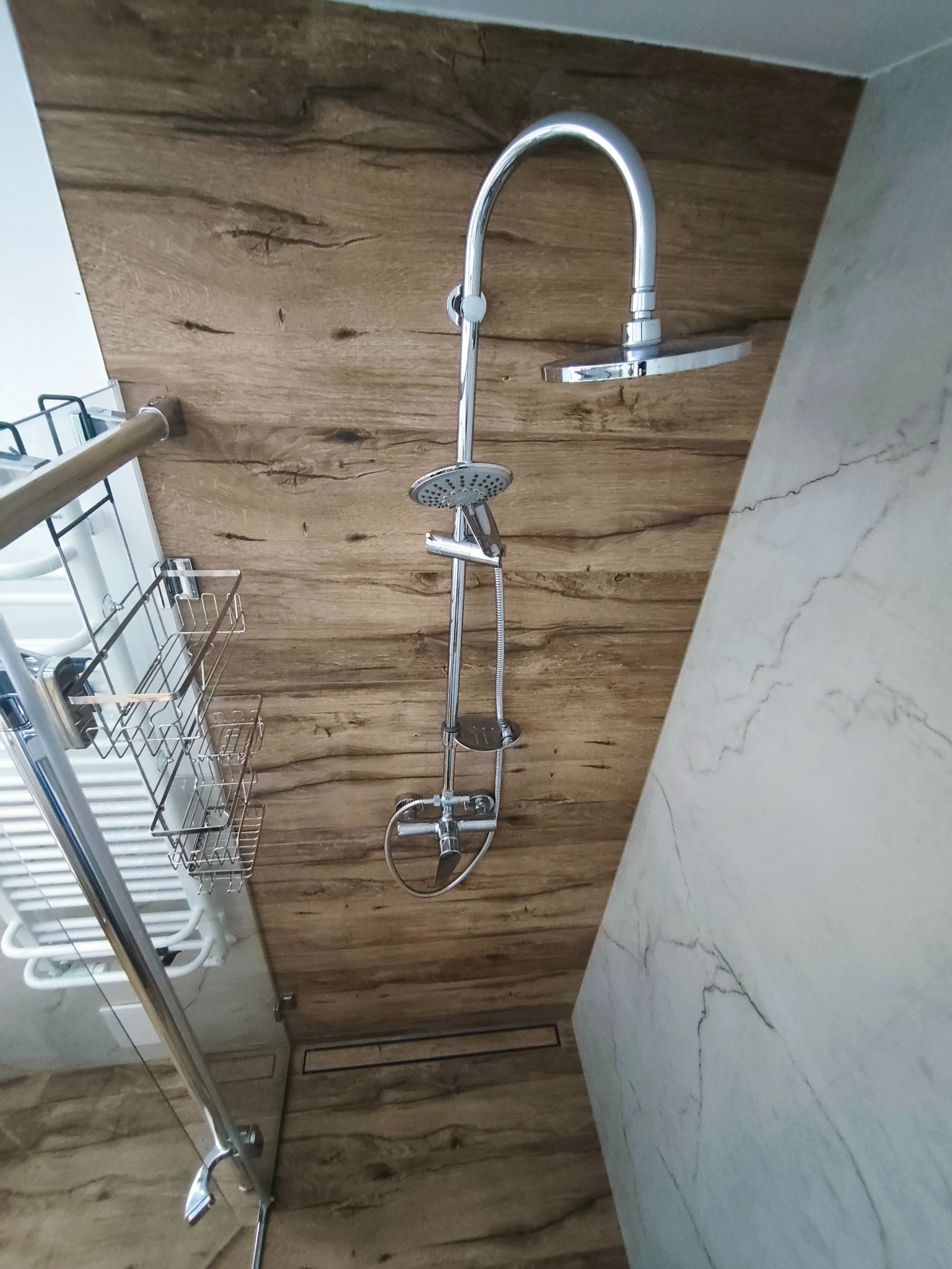 Wykończona kabina prysznicowa z chromowaną armaturą, drewnopodobnymi płytkami ściennymi i marmurową obudową. Widoczny odpływ liniowy i półki na kosmetyki.