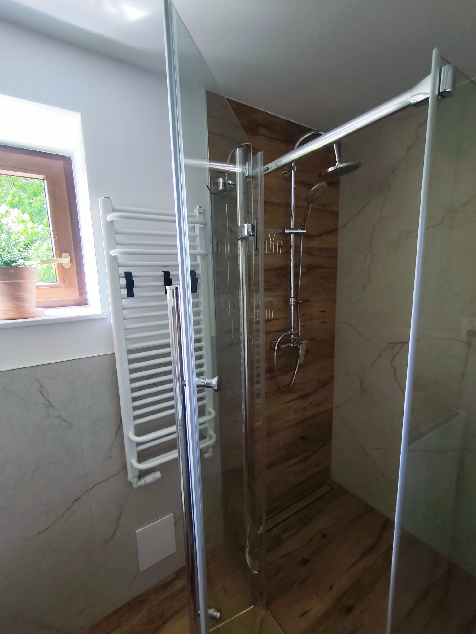 Nowoczesna kabina prysznicowa z chromowanymi okuciami, drewnopodobne płytki, grzejnik drabinkowy i okno z widokiem na zieleń.