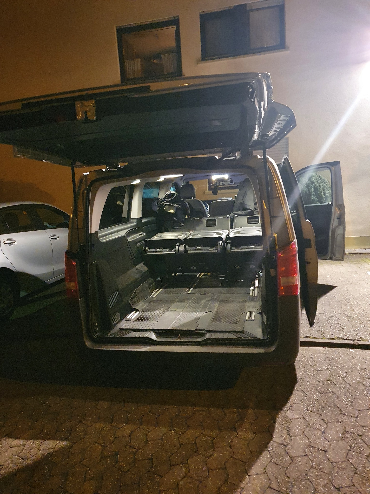 Wnętrze czarnego vana z otwartą klapą bagażnika, widoczne trzy rzędy siedzeń, bagaże i osobiste rzeczy pasażerów, na zewnątrz zaparkowany biały samochód osobowy.