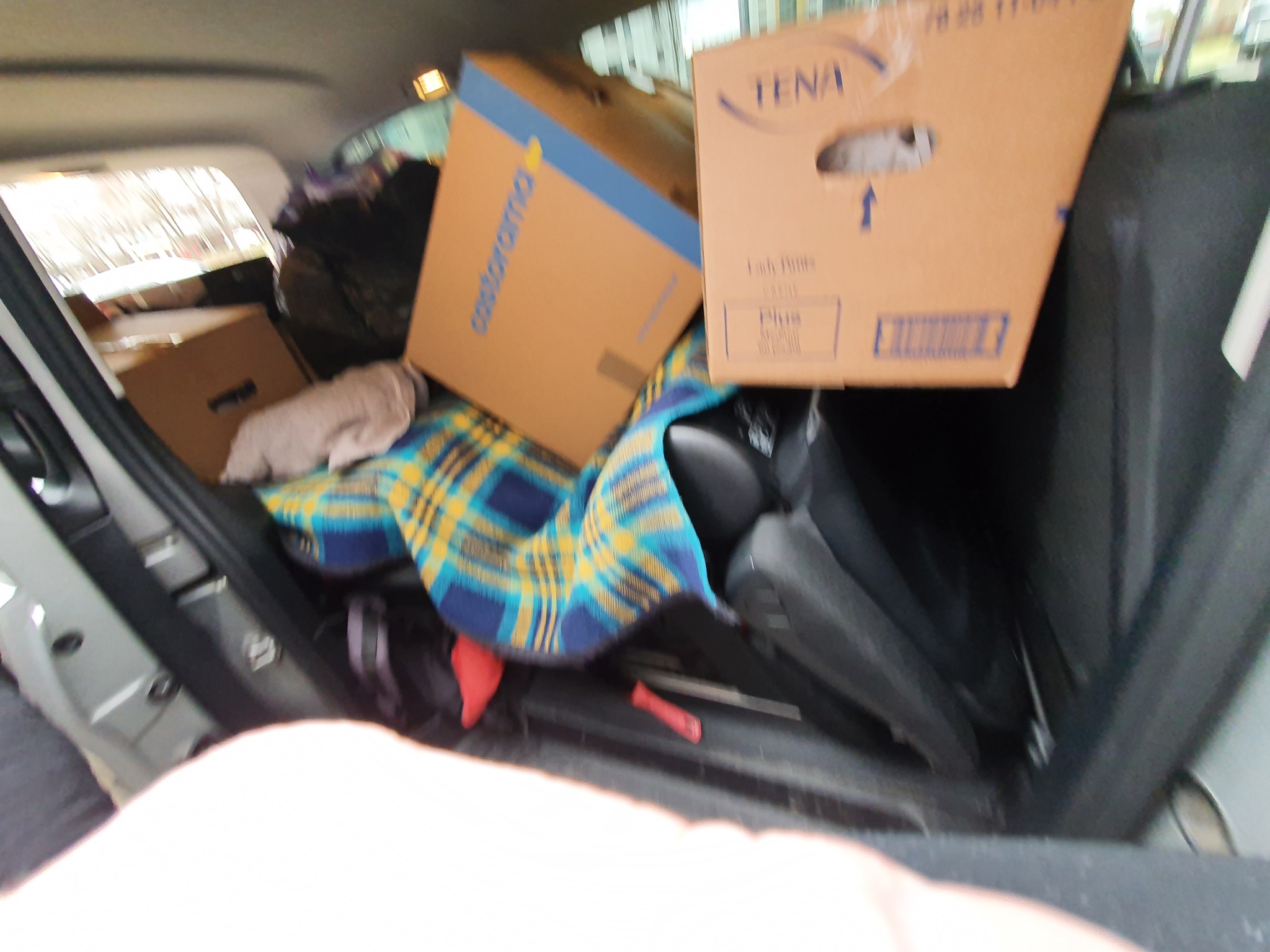 Wnętrze bagażnika samochodu osobowego załadowane kartonami Castorama i Tena, zabezpieczone niebiesko-żółtą pledem w kratę, widoczne przedmioty osobiste i częściowo złożone tylne siedzenia.