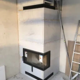 Wnętrze w trakcie wykańczania z kominkiem obudowanym płytami gipsowo-kartonowymi, widoczna drabina i poziomica, ściany surowe, brak wykończenia.