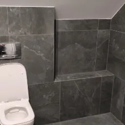 Nowoczesna toaleta z szarymi płytkami imitującymi kamień, podwieszana miska WC z chromowanym przyciskiem spłukującym i półką w zabudowie.