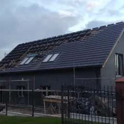 Dom w trakcie budowy z częściowo położonym, ciemnym dachem z dachówki ceramicznej i oknami dachowymi, ściany zewnętrzne ocieplone styropianem, widoczne rusztowania i materiały budowlane za...