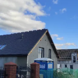 Dwa domy w trakcie budowy, jeden z ciemną dachówką i oknami dachowymi, drugi z widoczną konstrukcją dachu, oba z ocieplonymi ścianami, na tle błękitnego nieba z chmurami.