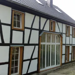 Elewacja domu w stylu szachulcowym, z nowymi oknami z drewnianą ramą, częściowo jeszcze nieotynkowana, z widoczną konstrukcją drewnianą.