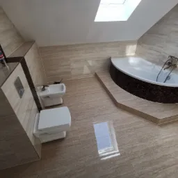 Ukończona łazienka na poddaszu z beżowymi płytkami imitującymi drewno, białą armaturą (toaleta, bidet) i narożną wanną obłożoną mozaiką; widoczne okno dachowe i akcesoria na półce.