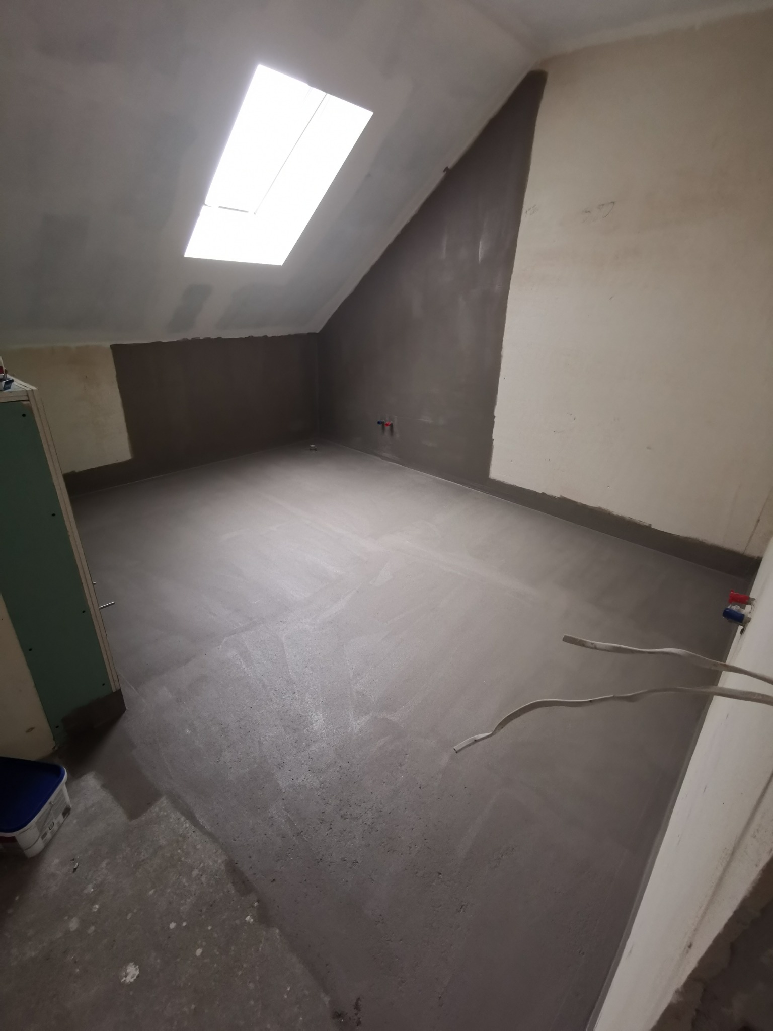 Poddasze w trakcie prac wykończeniowych: ściany częściowo pokryte gładzią, sufit z oknem dachowym, widoczne przewody elektryczne i wiadro z farbą.