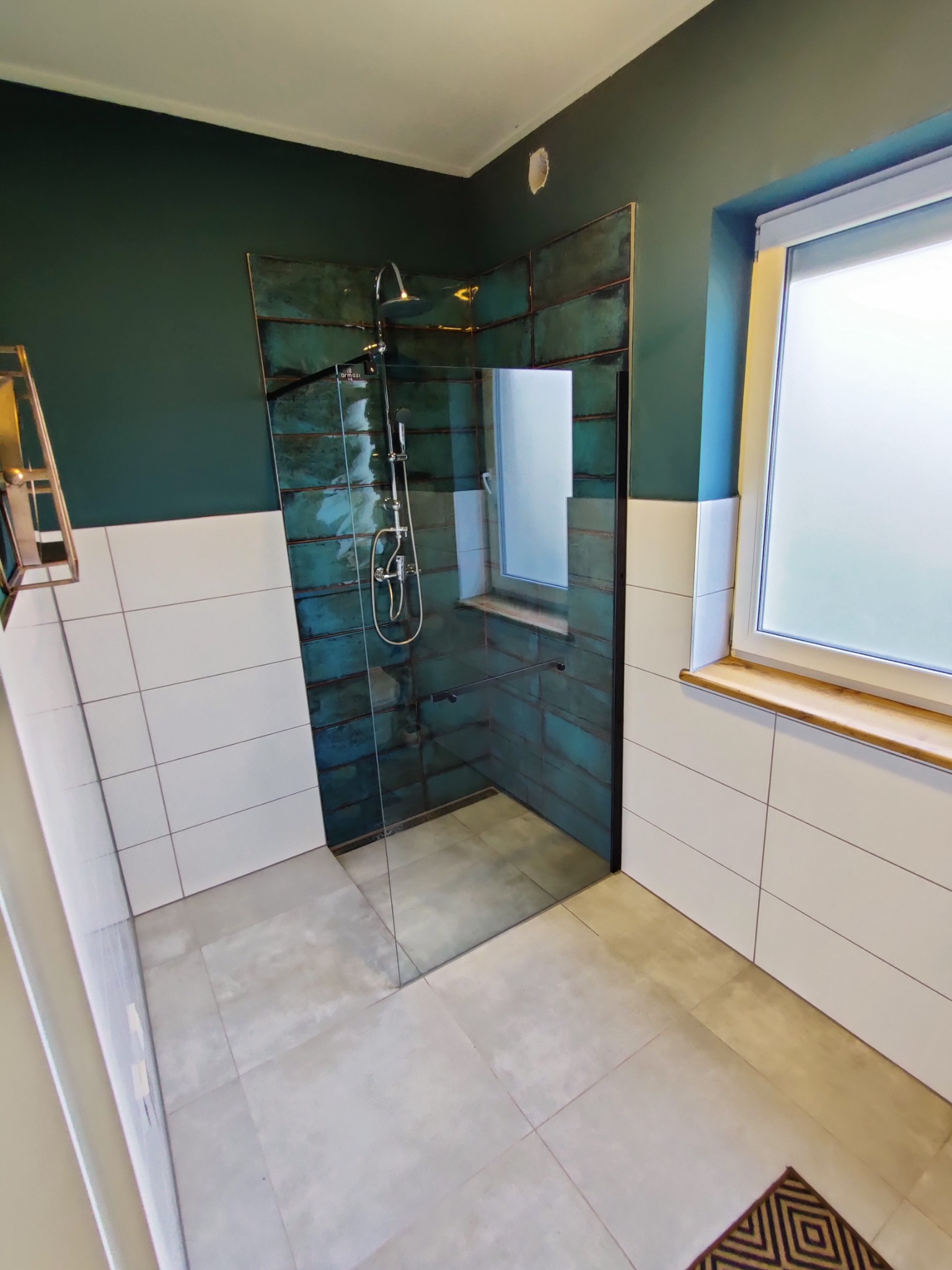 Nowoczesna łazienka z kabiną prysznicową typu walk-in, wykończona turkusowymi płytkami imitującymi patynowany metal w strefie prysznica, kontrastująca z białymi płytkami na pozostałej części ścian...