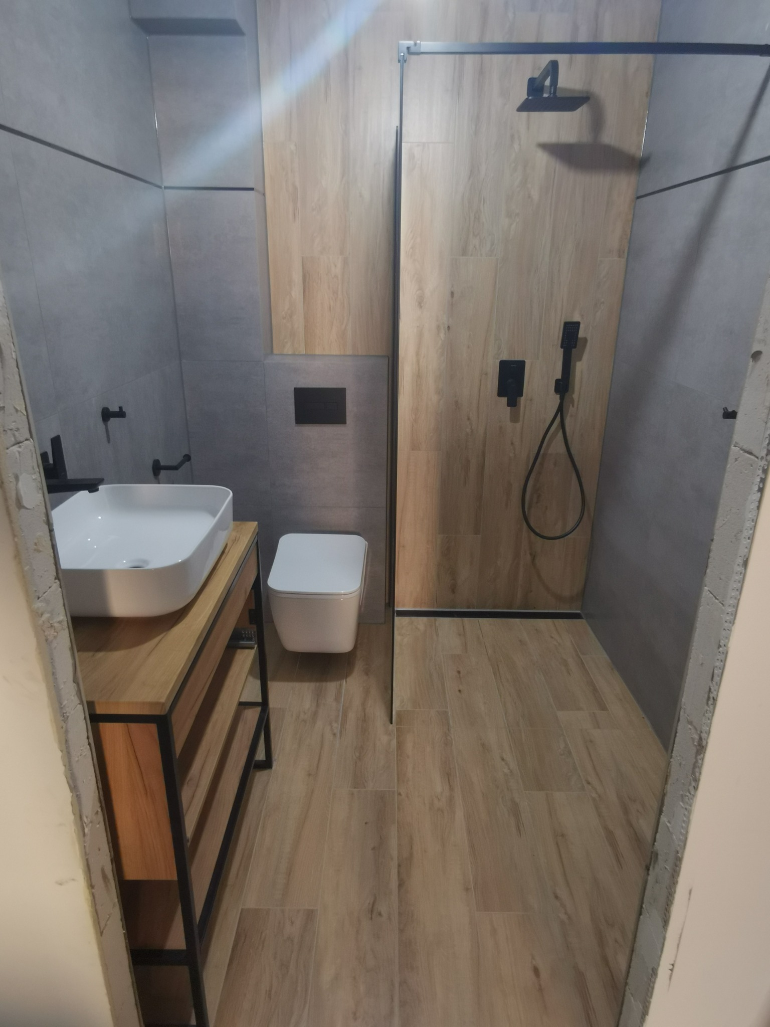 Nowoczesna łazienka z drewnopodobnymi płytkami w kabinie prysznicowej, szare płytki na ścianach, biała umywalka nablatowa na drewnianej szafce z czarnymi metalowymi elementami oraz wisząca toaleta.