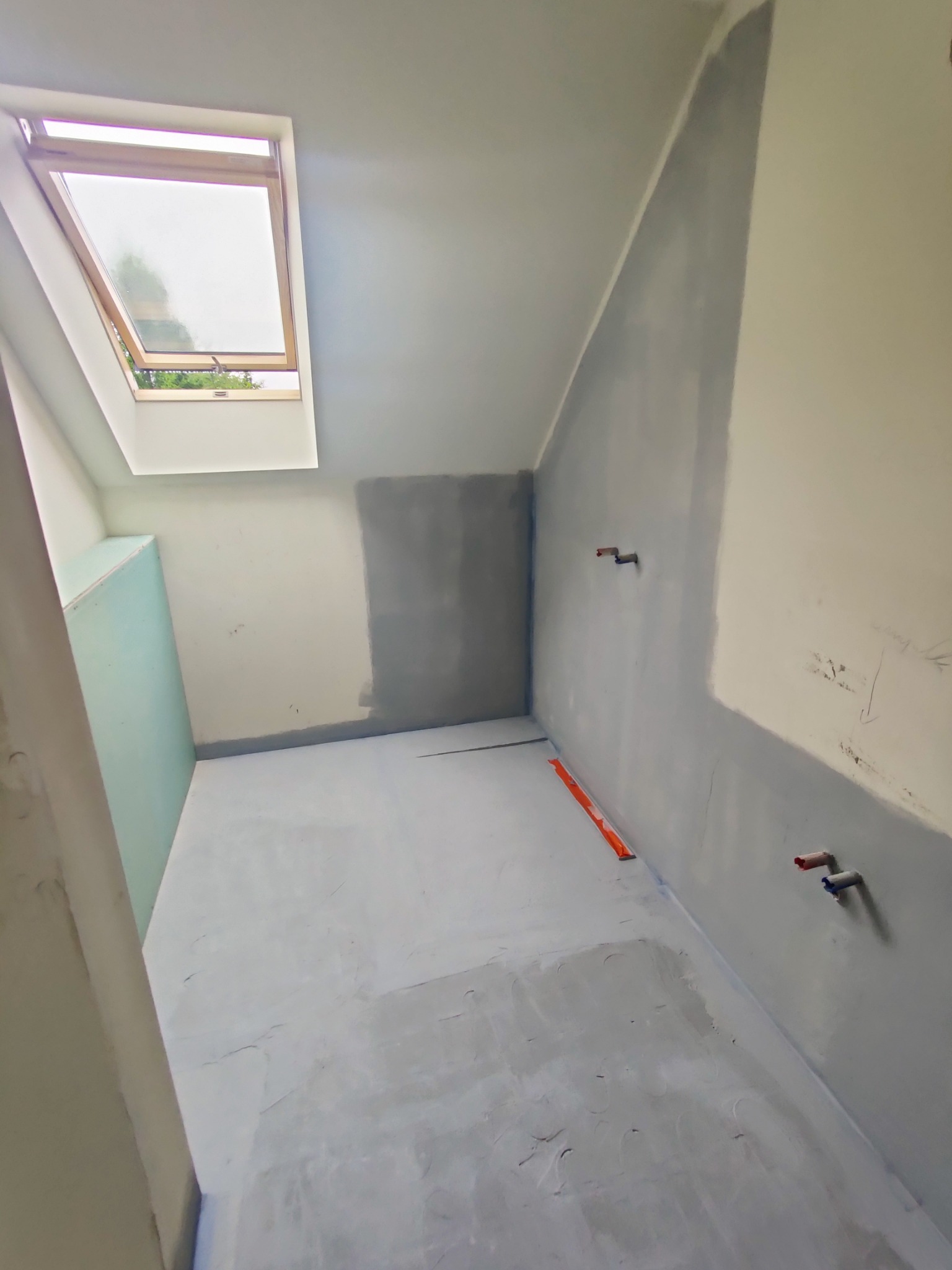 Poddasze w trakcie remontu: częściowo zagruntowane ściany, wylewka na podłodze, okno dachowe, poziomica na podłodze, widoczne wystające rury instalacji wodnej.