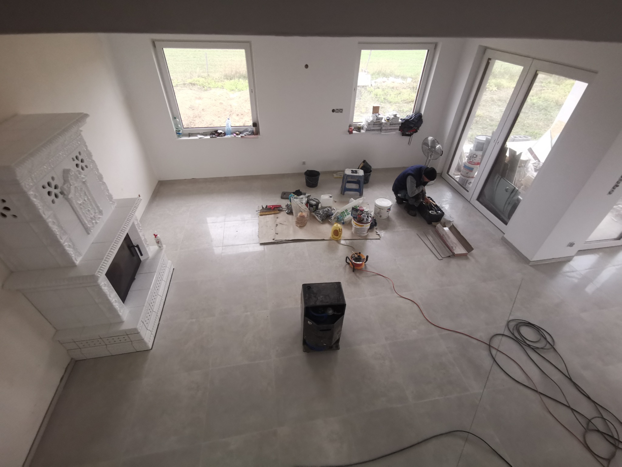 Wnętrze w trakcie wykańczania: ułożona jasna podłoga z płytek, biały ozdobny kominek, okna dachowe i dwuskrzydłowe drzwi balkonowe, widoczne narzędzia i materiały budowlane oraz pracownik w trakcie...