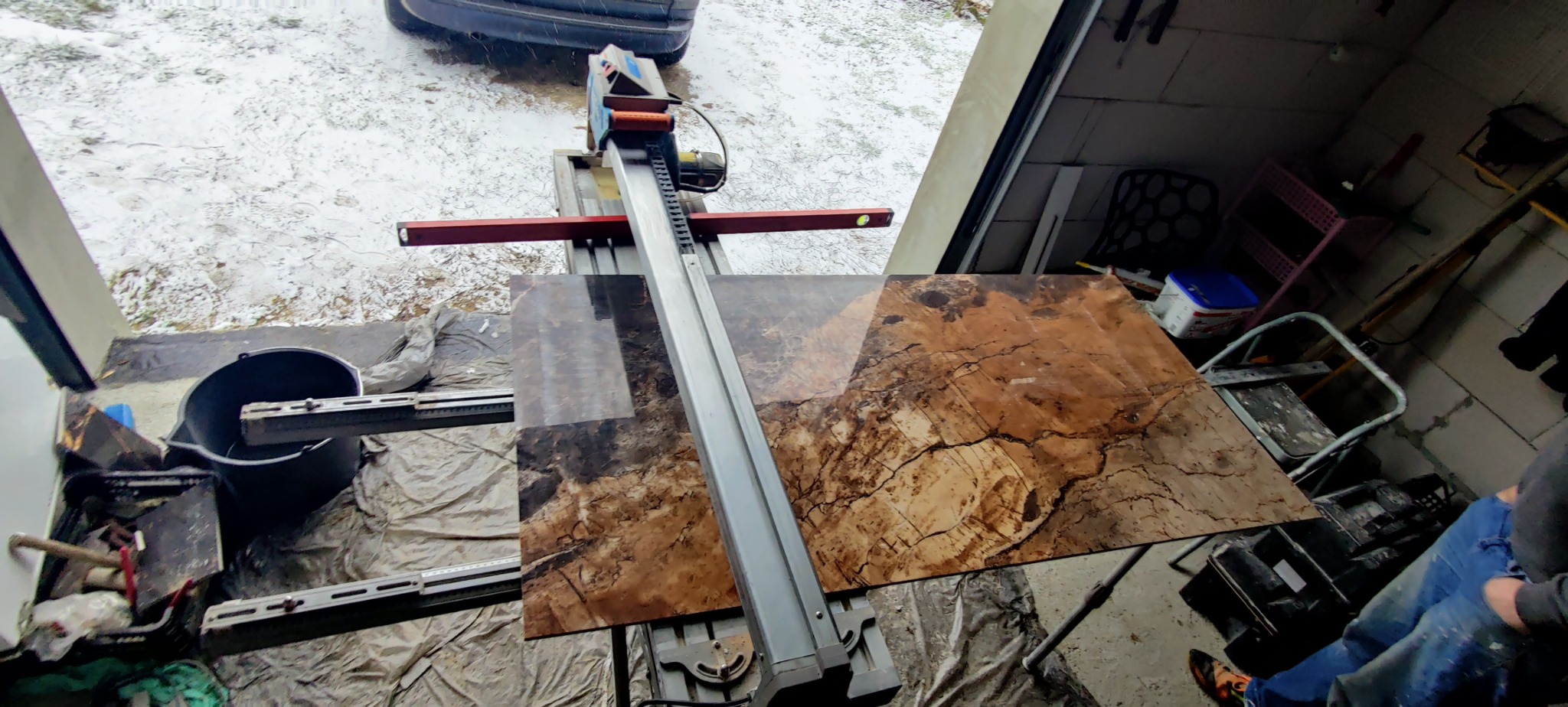 Cięcie brązowej płytki imitującej marmur na mokro przecinarce w garażu, widoczny fragment osoby w jeansach i narzędzia.