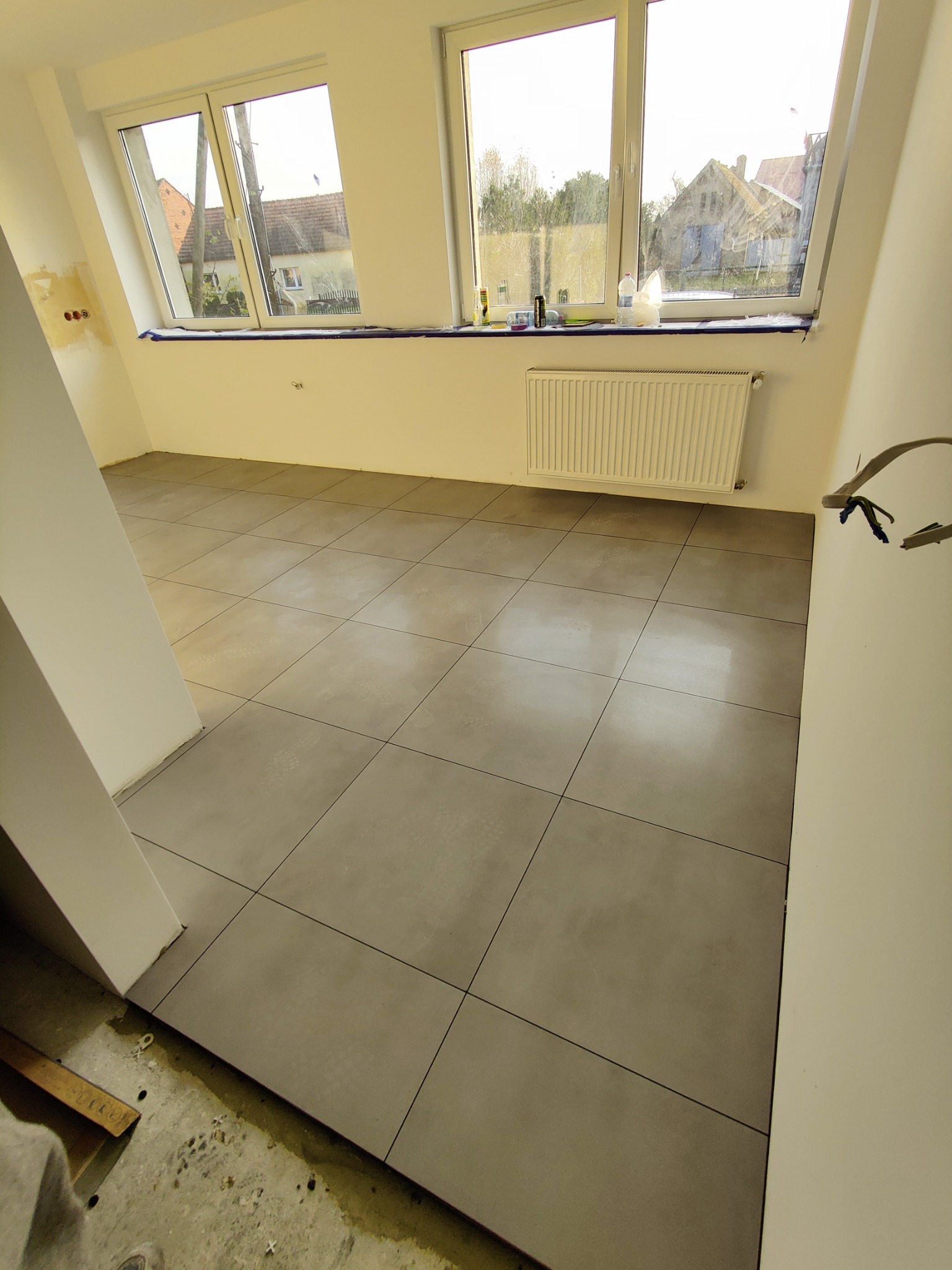 Świeżo ułożone, szare płytki podłogowe w jasnym pomieszczeniu z dwoma oknami, widoczne niedokończone prace przy listwach przypodłogowych i wystające przewody elektryczne.