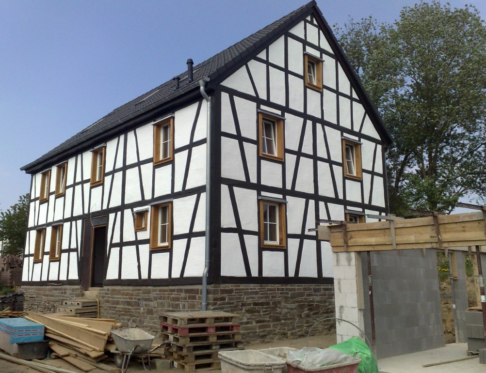 Dwukondygnacyjny dom w stylu szachulcowym z białymi ścianami i ciemnymi belkami, widoczny w trakcie budowy lub remontu, z elementami konstrukcyjnymi i materiałami budowlanymi na pierwszym planie.