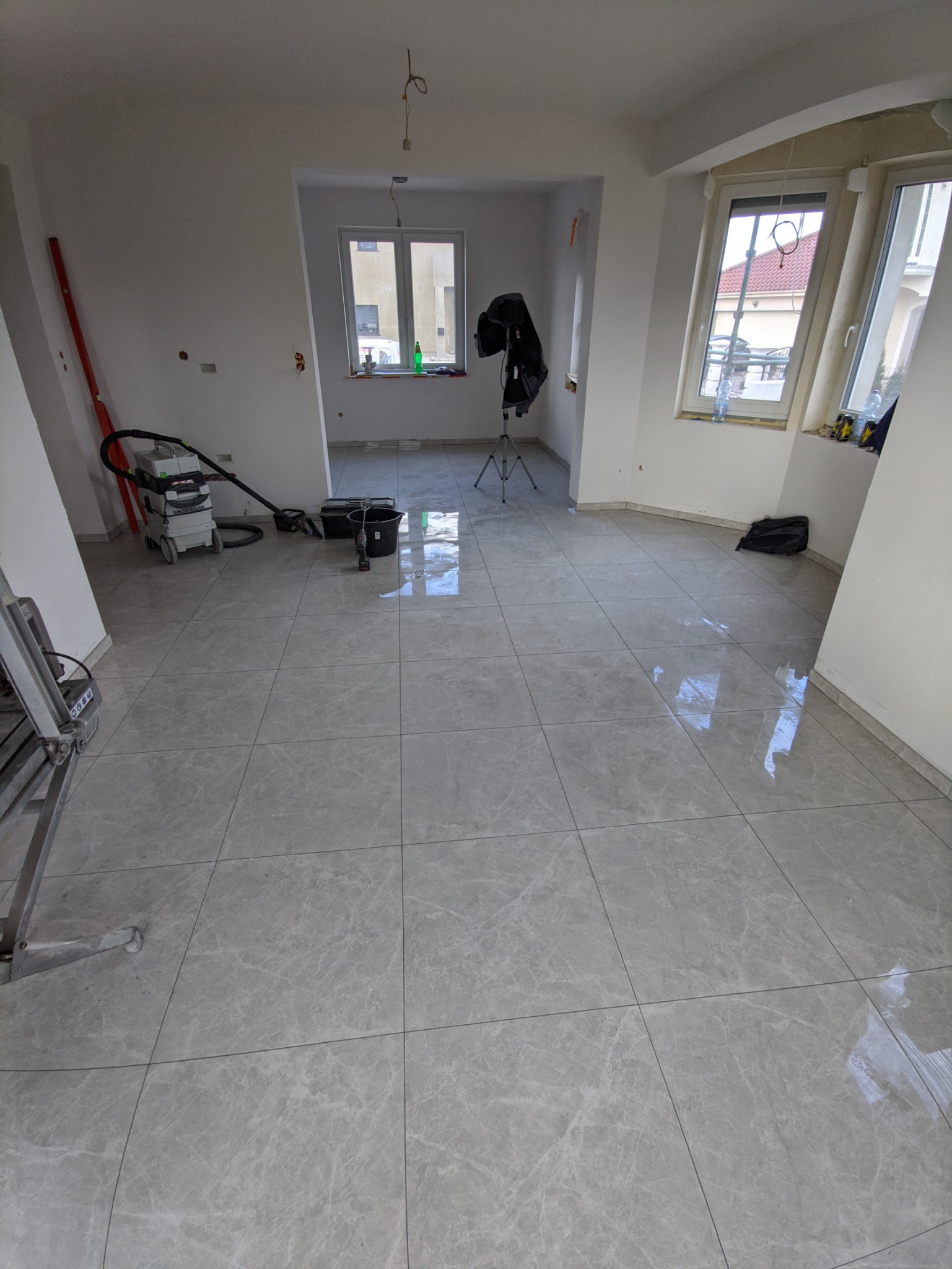 Wykończone wnętrze z szarymi płytkami podłogowymi imitującymi marmur, widoczny sprzęt budowlany, okna z widokiem na budynki.