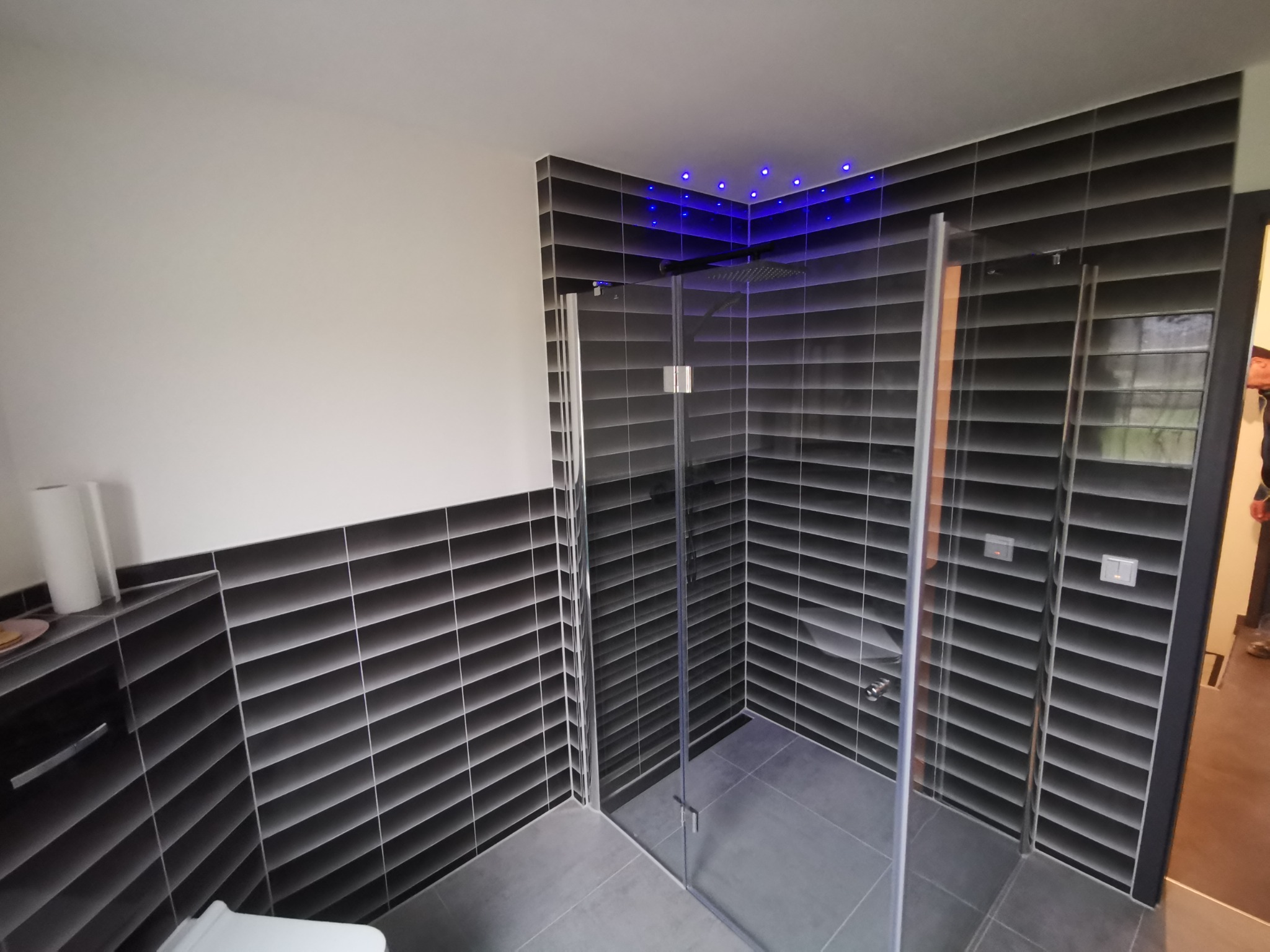 Nowoczesna kabina prysznicowa z czarnymi, poziomymi płytkami, podświetleniem LED w suficie i szklanymi drzwiami, widok z zewnątrz.