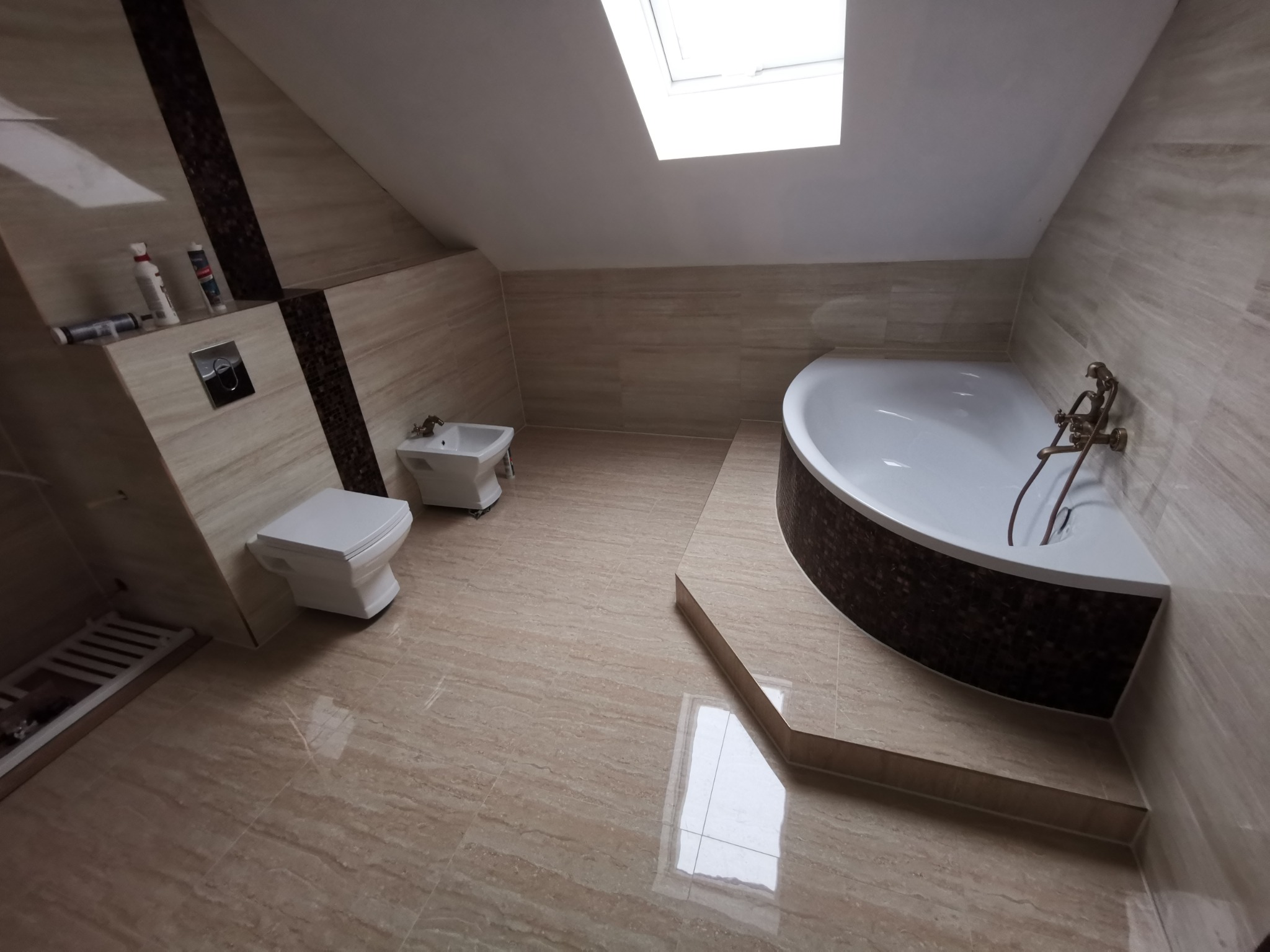 Wykończona łazienka na poddaszu ze skośnym sufitem, z beżowymi płytkami imitującymi marmur na podłodze i ścianach, białą armaturą (toaleta, bidet, wanna narożna z mozaiką) i oknem dachowym...