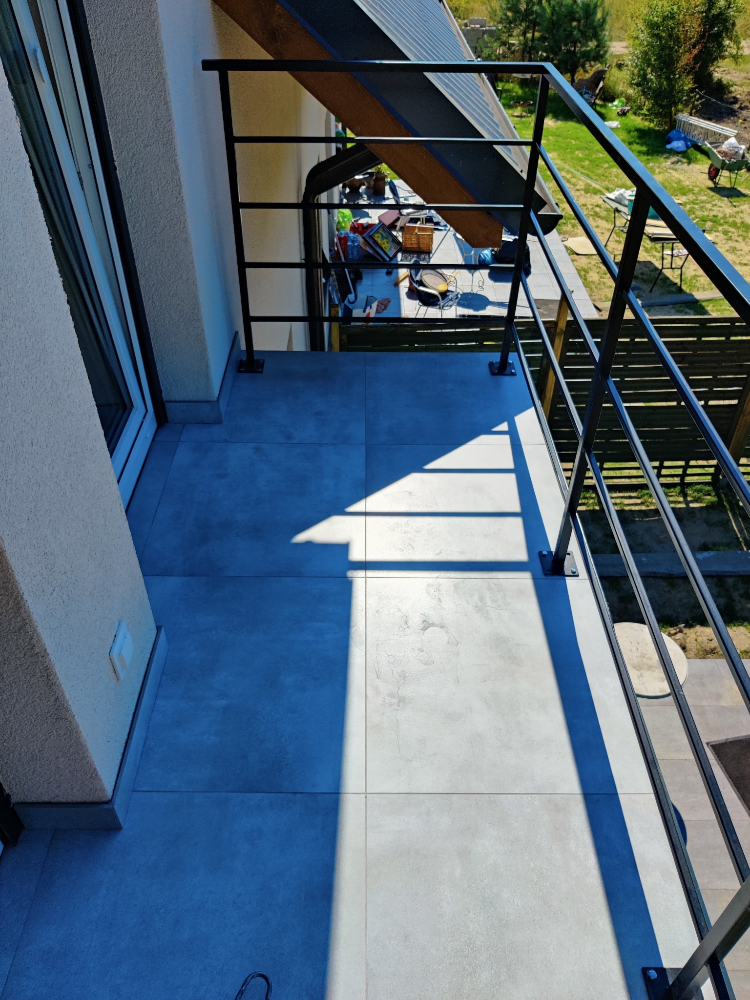 Szare płytki na balkonie z czarną metalową balustradą, widok z góry, cień balustrady na podłodze, widok na ogród z elementami wyposażenia.