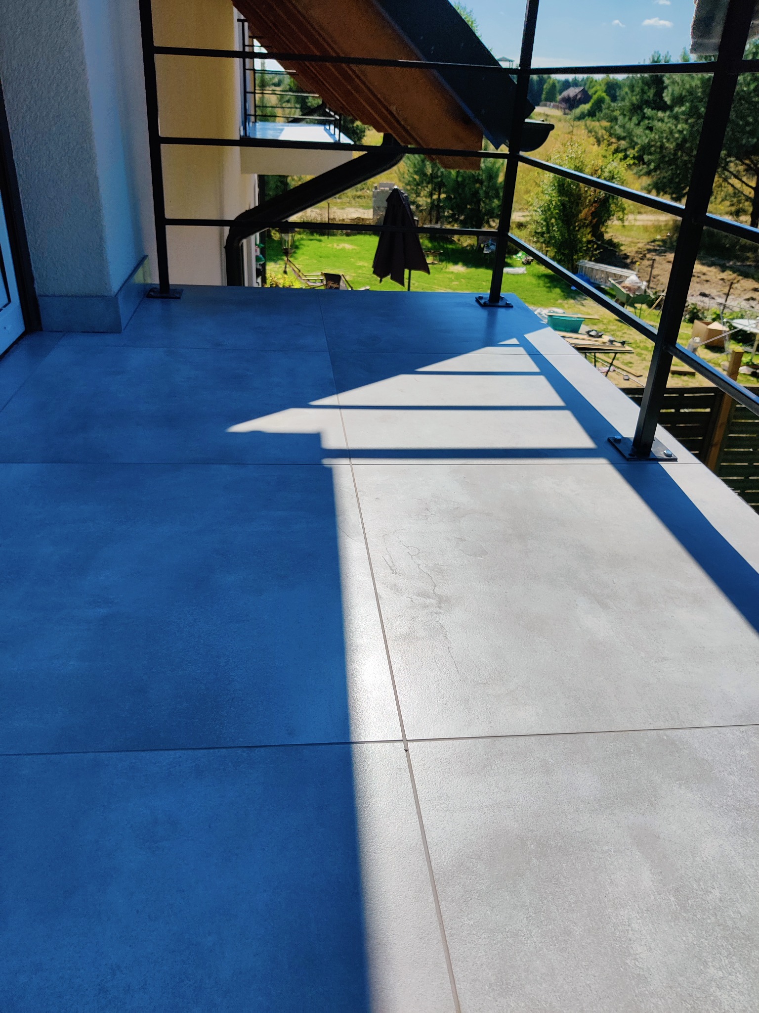 Szare płytki na balkonie z widocznymi cieniami, czarne metalowe barierki, zieleń w tle.