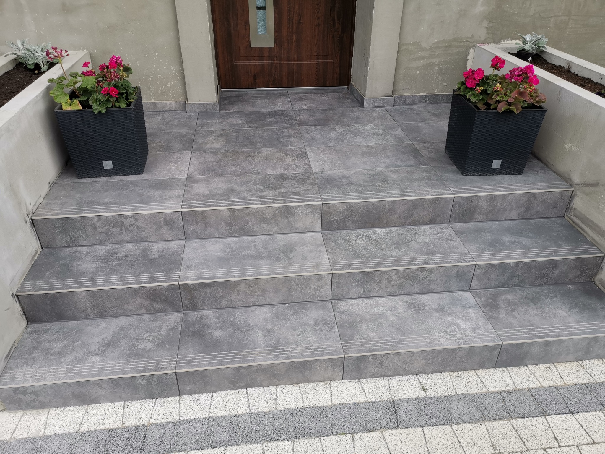 Schody wejściowe wyłożone szarymi płytkami z trzema antypoślizgowymi paskami na każdym stopniu, ozdobione po bokach czarnymi donicami z czerwonymi kwiatami, prowadzące do drewnianych drzwi.