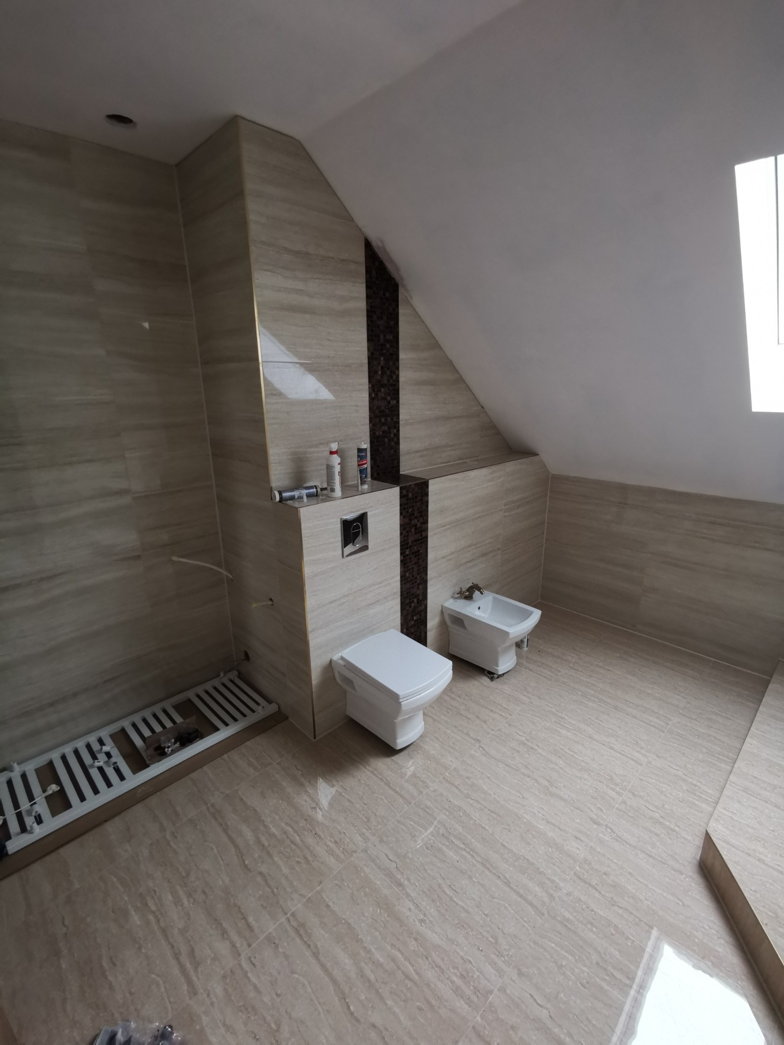 Wykończona łazienka na poddaszu z beżowymi płytkami imitującymi drewno, wisząca toaleta i bidet, z widocznymi akcesoriami montażowymi i narzędziami.