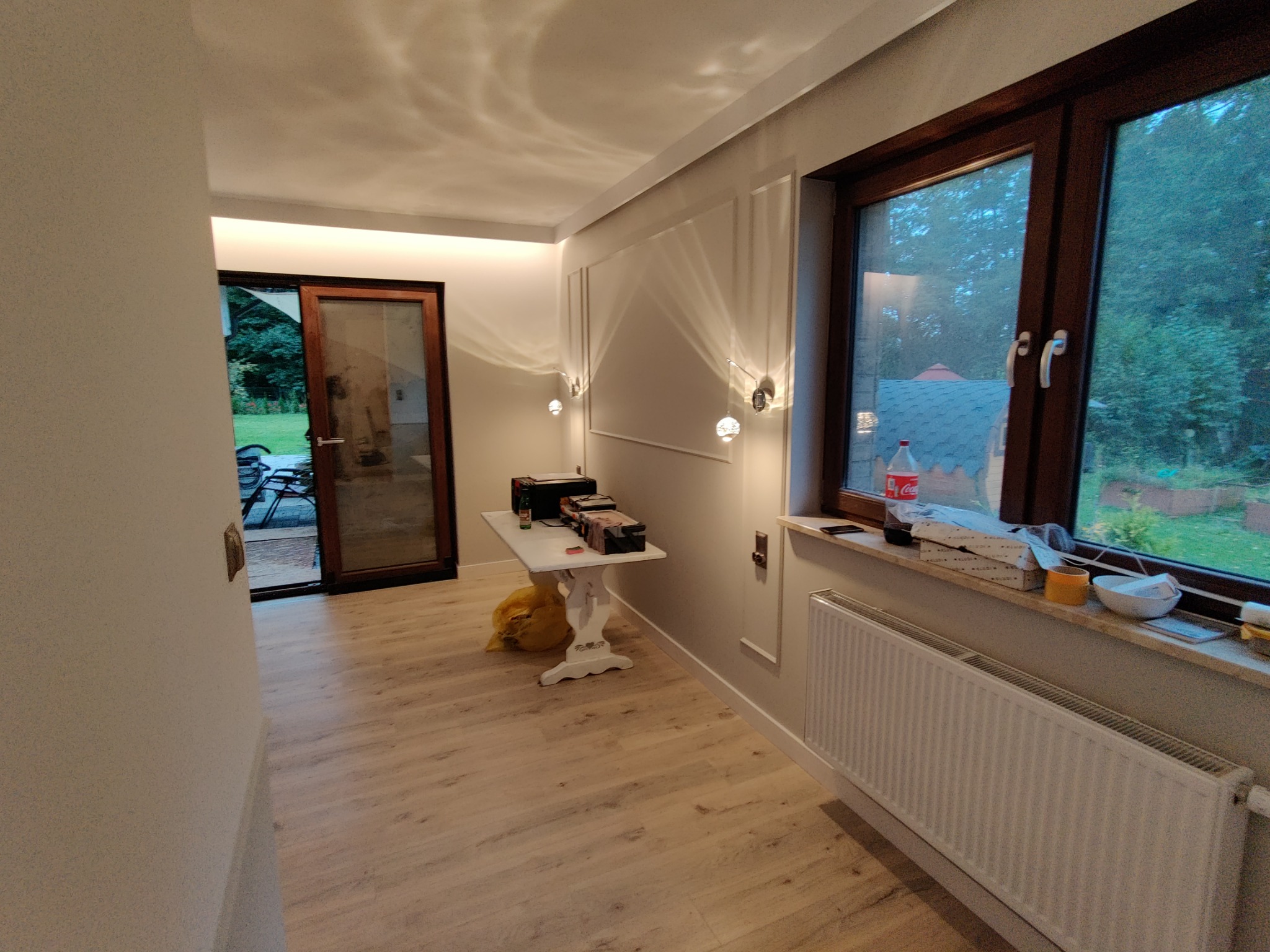 Jasne wnętrze po remoncie: parkiet, grzejnik pod oknem, dekoracyjne listwy na ścianie, oświetlenie punktowe i biały stolik z akcesoriami.