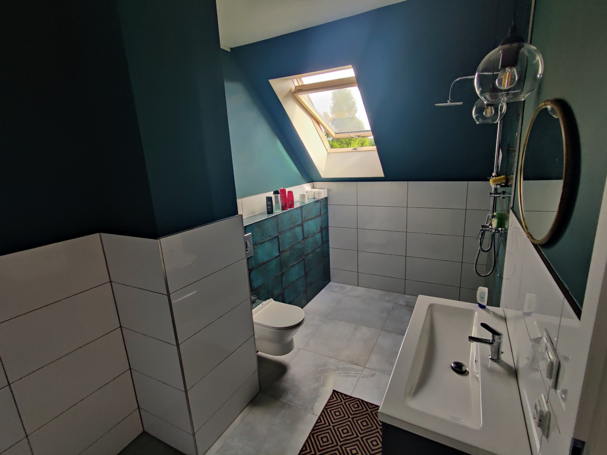 Nowoczesna łazienka na poddaszu z oknem dachowym, biało-turkusowymi płytkami, ciemną ścianą i geometrycznym dywanikiem.