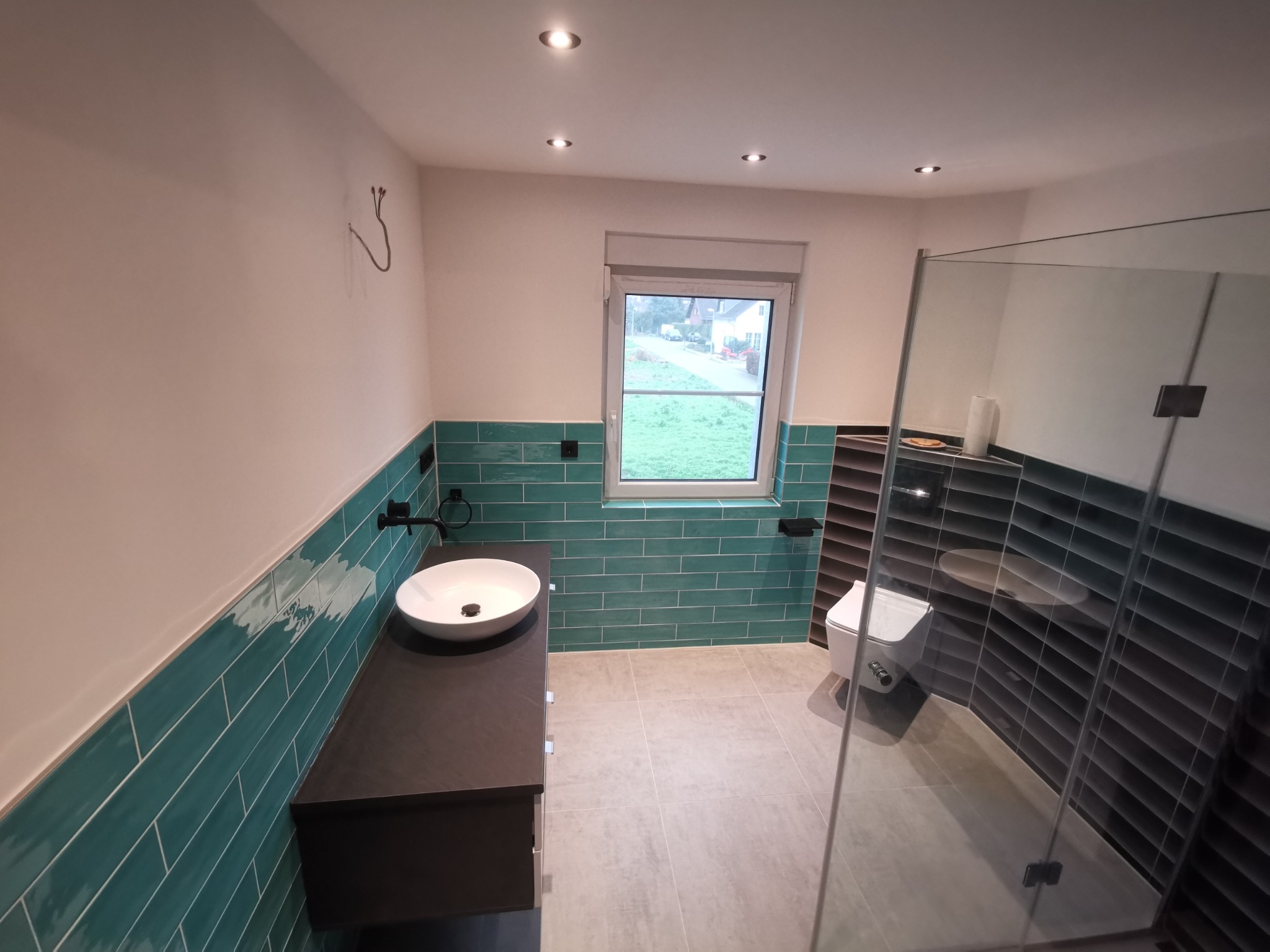 Nowoczesna łazienka z turkusowymi płytkami ściennymi ułożonymi w cegiełkę, ciemnym blatem pod umywalkę nablatową, szarą podłogą i kabiną prysznicową ze szkła.