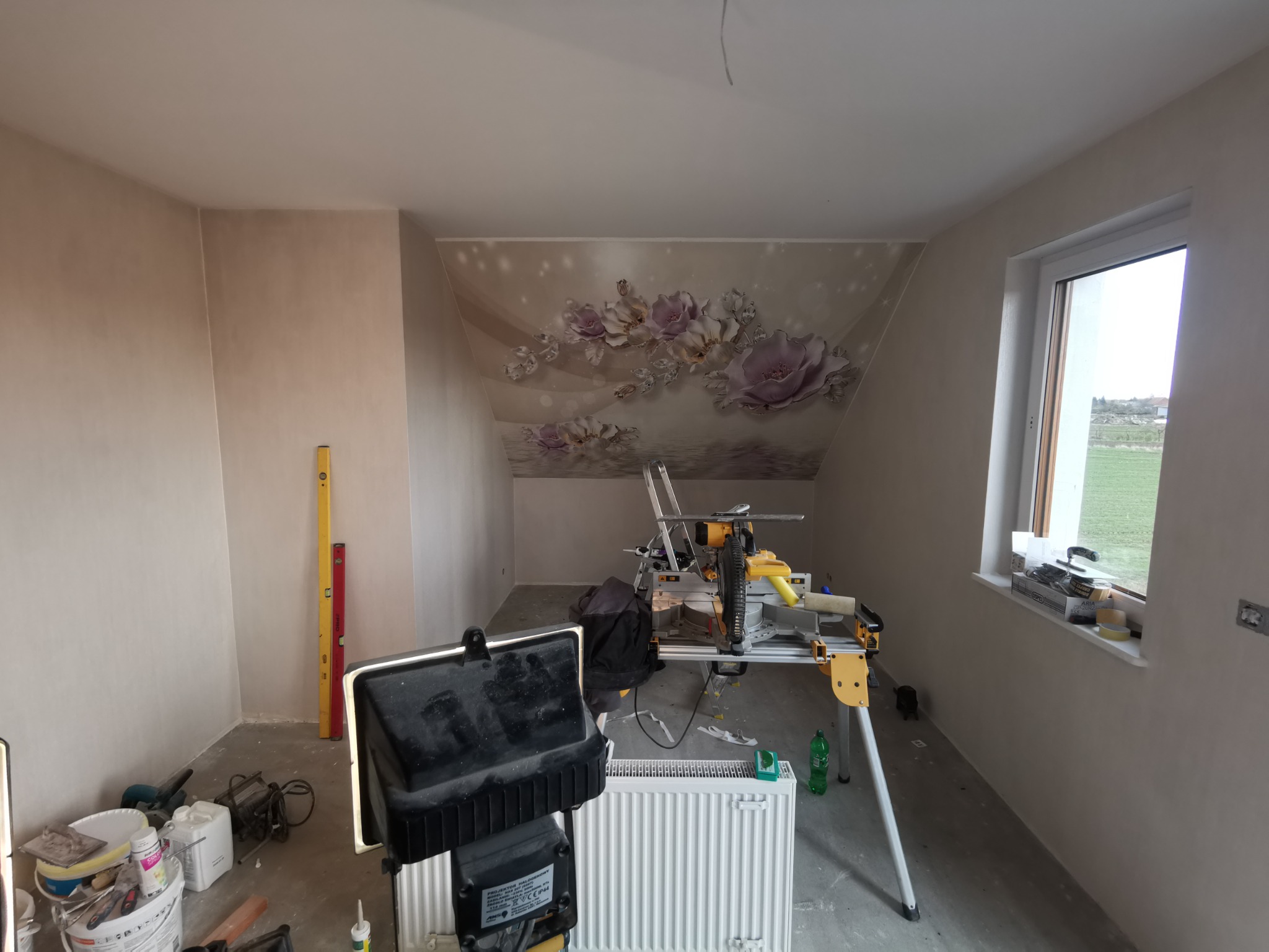 Wnętrze pokoju w trakcie wykańczania, ściany po nałożeniu gładzi, widoczny sprzęt do cięcia płyt, poziomice, lampa budowlana i grzejnik, na ścianie dekoracyjny wzór kwiatowy.