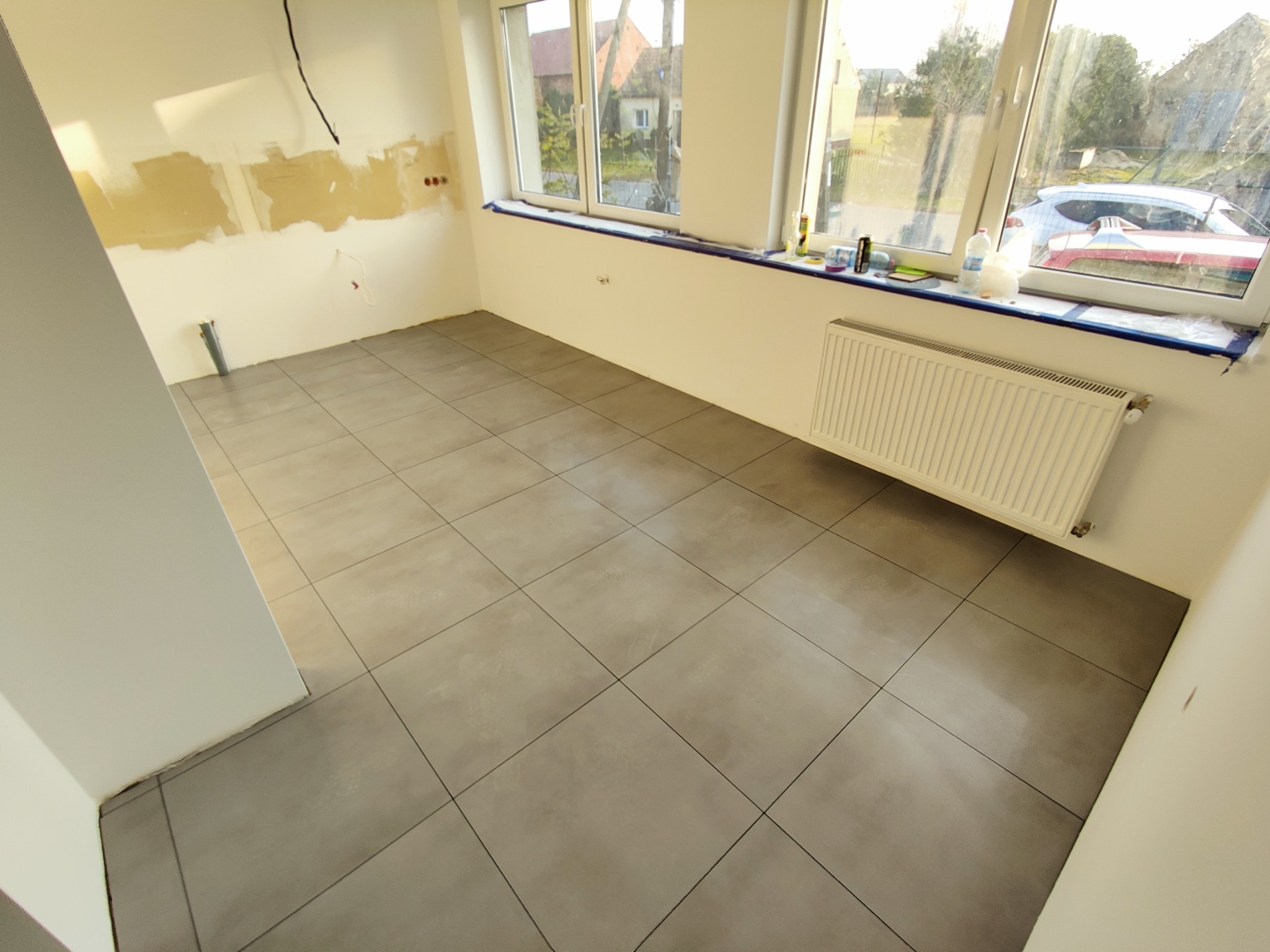 Świeżo ułożone szare płytki podłogowe w jasnym pomieszczeniu z widocznymi śladami prac remontowych na ścianach, grzejnikiem i oknem z widokiem na zewnątrz.