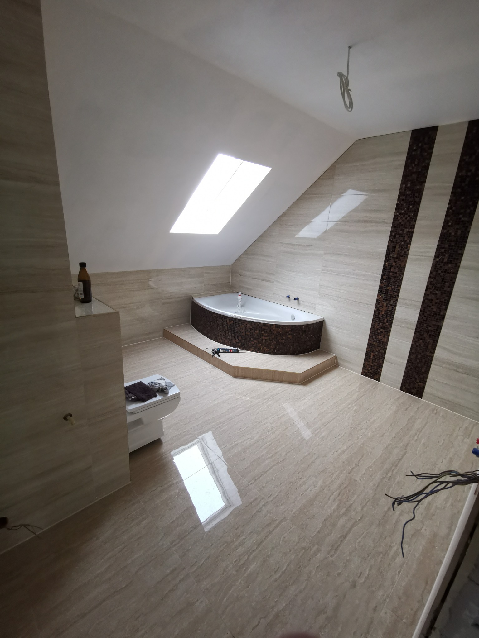 Wykończona łazienka na poddaszu z wanną obudowaną mozaiką, beżowe płytki imitujące marmur na ścianach i podłodze, okno dachowe w skosie, widoczne niedokończone podłączenia elektryczne.