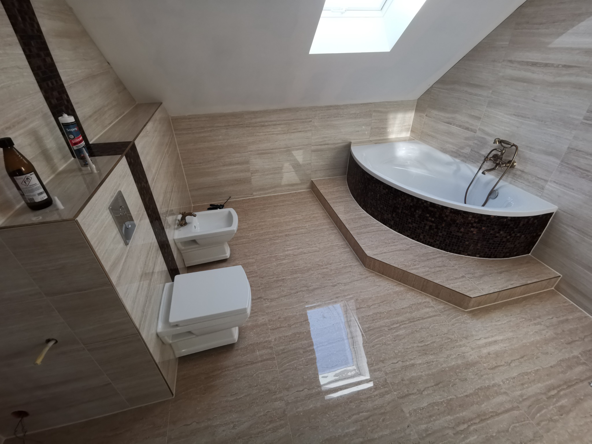 Ukończona łazienka na poddaszu z beżowymi płytkami imitującymi drewno, białą armaturą (toaleta, bidet) i narożną wanną obłożoną mozaiką; widoczne okno dachowe i akcesoria na półce.