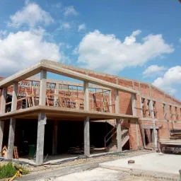 Dwukondygnacyjny budynek żelbetowy w trakcie budowy, z widocznymi czerwonymi cegłami, betonowymi słupami i stropami, drabiny opierające się o ściany, na zewnątrz zaparkowany samochód dostawczy...