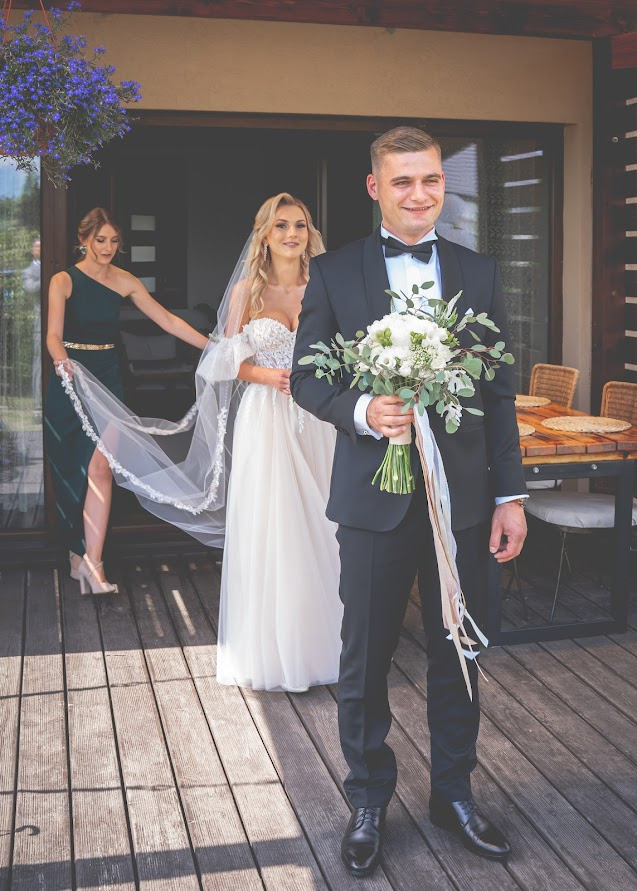 Uśmiechnięty pan młody w czarnym smokingu, trzymający bukiet białych kwiatów i zielonych liści, obok panny młodej w białej sukni z welonem i druhny w zielonej sukni, wychodzący z budynku...