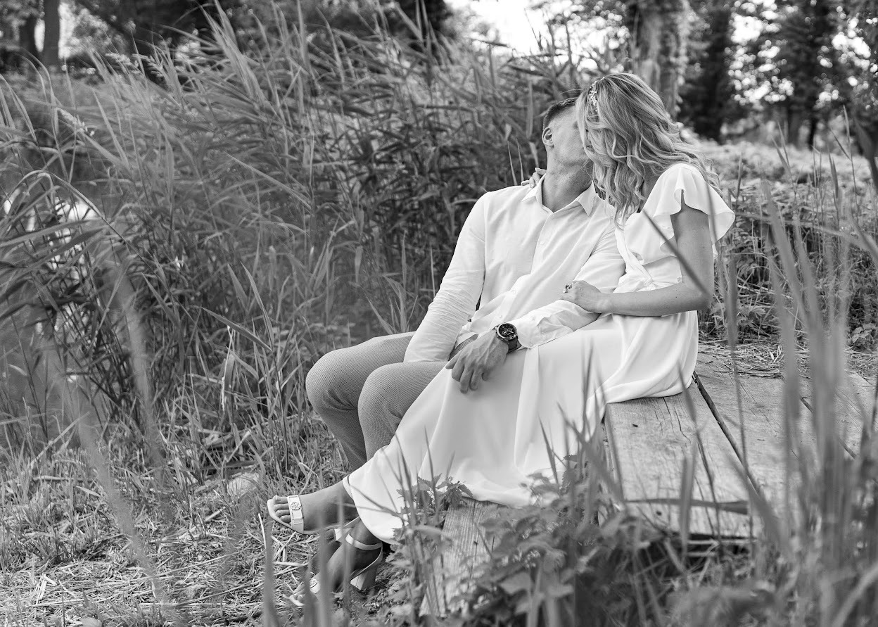 Czarno-białe zdjęcie zakochanej pary siedzącej na drewnianym pomoście wśród traw i trzcin, delikatny pocałunek, ona w zwiewnej białej sukience i sandałach na obcasie, on w koszuli i spodniach,...