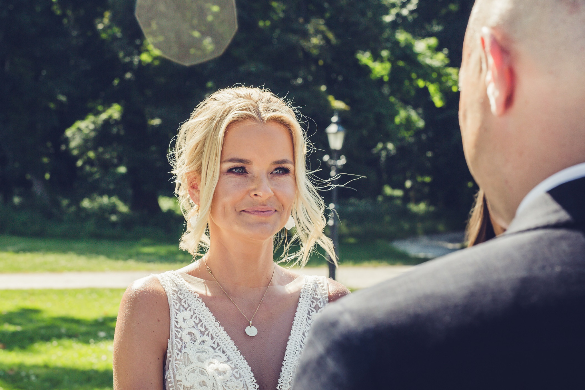 Panna młoda w koronkowej sukni spogląda z uśmiechem na pana młodego podczas sesji narzeczeńskiej w plenerze z parkiem w tle.