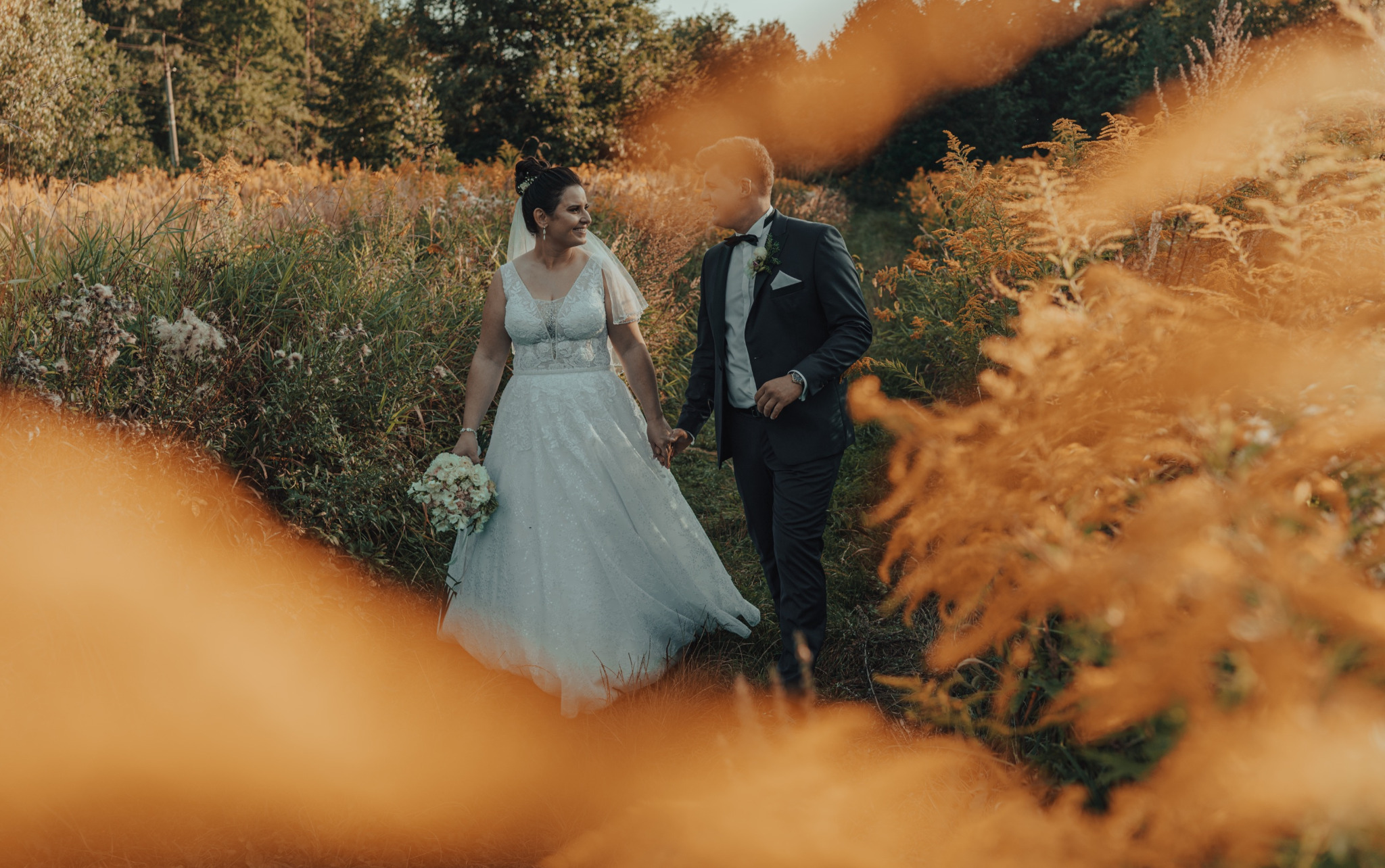 Para młoda, on w ciemnym garniturze z muchą, ona w białej sukni z bukietem, spacerują po łące z wysoką trawą w ciepłym świetle zachodzącego słońca, patrząc na siebie z uśmiechem.