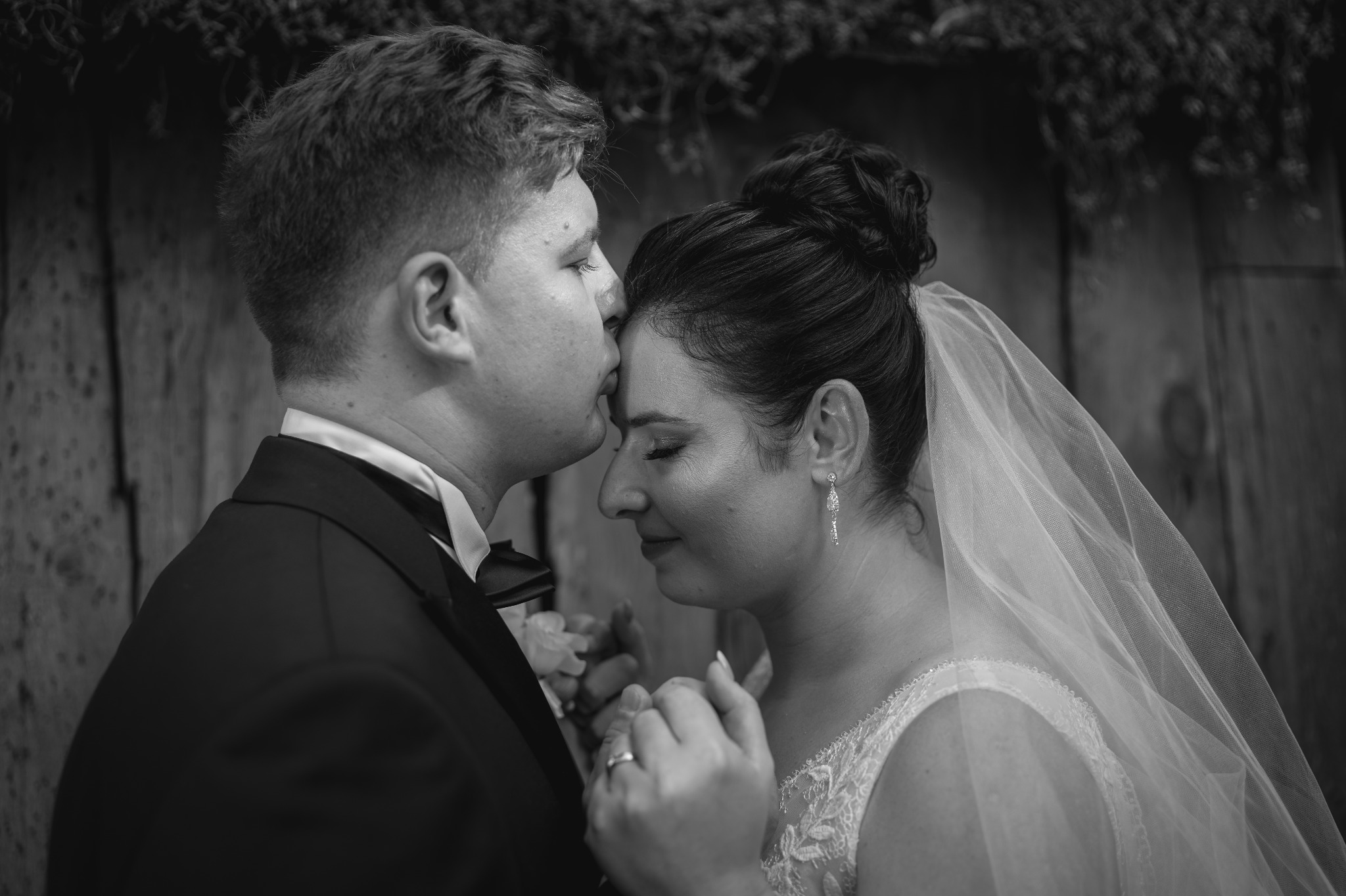 Czarno-białe zbliżenie nowożeńców: pan młody całuje pannę młodą w czoło, trzymając ją za ręce na tle drewnianej ściany z zielonymi akcentami.